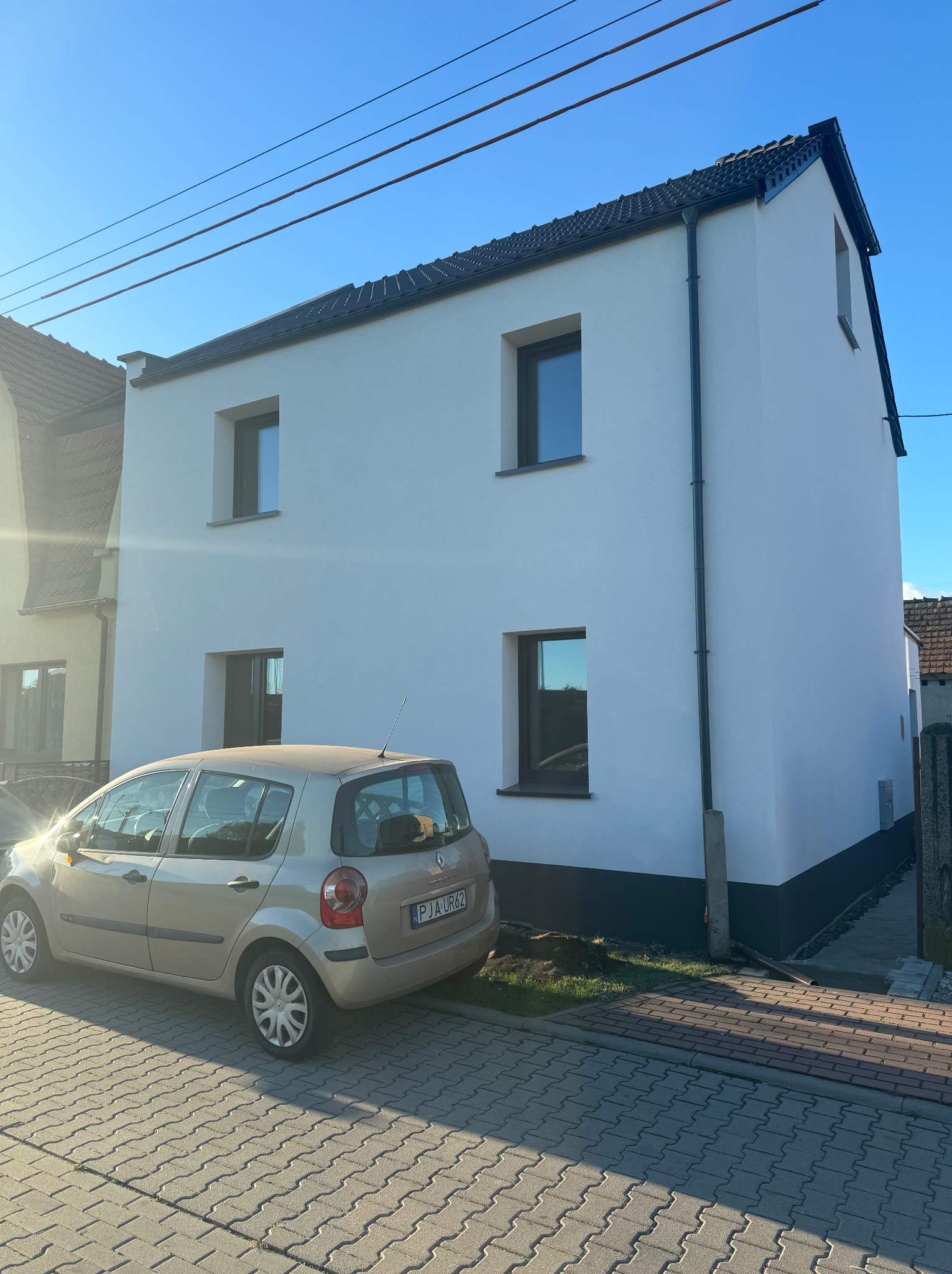 Biały dom z nową elewacją i ciemnymi oknami, zaparkowany samochód. Świeżo pomalowana fasada w słoneczny dzień. Widok na dom z chodnika.