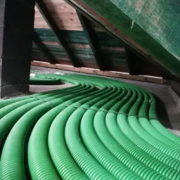 Zielone, karbowane rury wentylacyjne prowadzone wzdłuż poddasza z drewnianą konstrukcją dachu, zabezpieczone czarnymi opaskami.