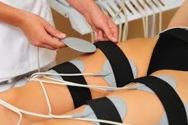 Elektrostymulacja ud z użyciem elektrod przymocowanych czarnymi pasami, widoczne dłonie osoby obsługującej urządzenie.