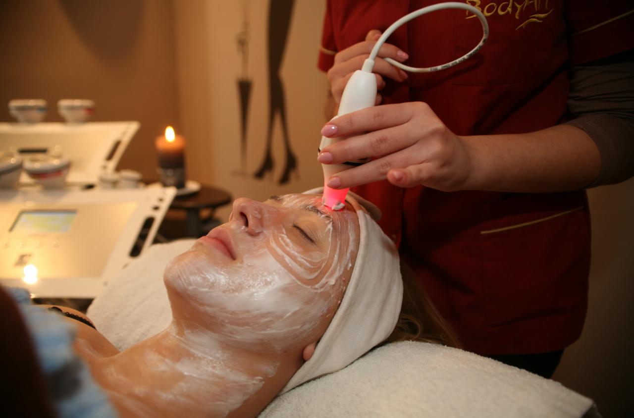 Kobieta z nałożoną maską na twarzy, poddawana zabiegowi z użyciem urządzenia emitującego czerwone światło, w tle zapalona świeca i sprzęt kosmetyczny.