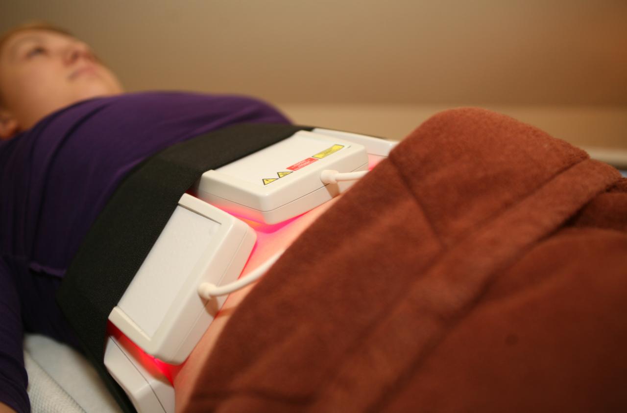 Klientka leżąca podczas zabiegu modelowania sylwetki z użyciem urządzenia z białymi aplikatorami emitującymi czerwone światło, przymocowanymi do ciała czarnym pasem.