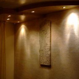 Usługi Remontowo Wykończeniowe - Pokój z dekoracyjną, teksturowaną ścianą w odcieniach beżu, podświetloną punktowymi lampami sufitowymi i zdobioną reliefem o motywach egipskich.