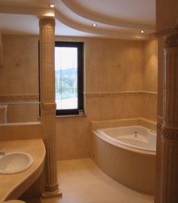 Wykończona łazienka w beżowych odcieniach z wanną narożną, umywalką wpuszczaną w blat i dekoracyjnymi kolumnami.