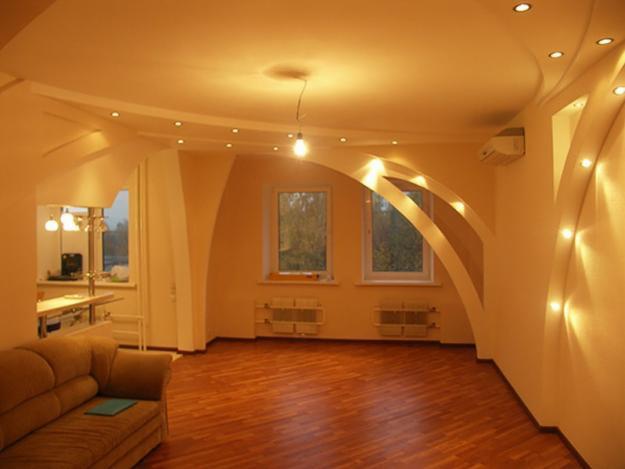 Jasne wnętrze z parkietem, dekoracyjnymi łukami i oświetleniem punktowym na suficie i ścianie; widoczne okna i fragment kuchni w tle.