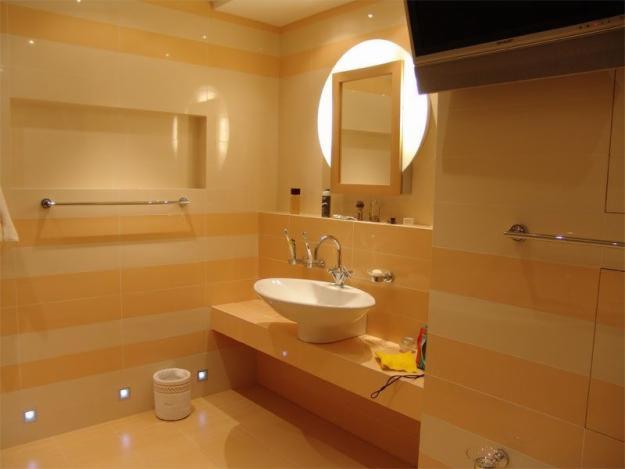 Elegancka łazienka z poziomymi pasami płytek w odcieniach beżu i pomarańczy, owalna umywalka nablatowa, okrągłe lustro z podświetleniem oraz wbudowane oświetlenie podłogowe.