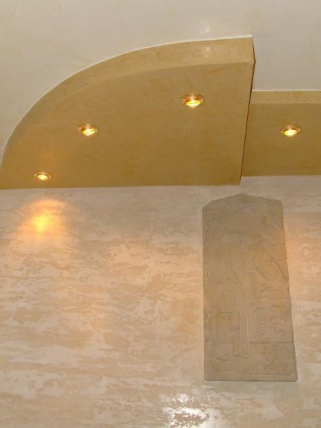 Beżowa ściana z efektem trawertynu, podświetlany sufit podwieszany z wbudowanymi punktami świetlnymi oraz dekoracyjna płaskorzeźba z motywami egipskimi.