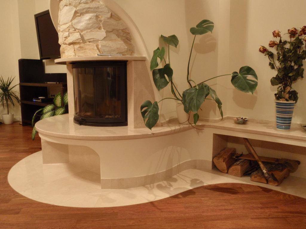 Ozdobny kominek z kamienną obudową i półokrągłym paleniskiem, drewno do kominka w niszy, drewniana podłoga, rośliny doniczkowe w tle, telewizor.