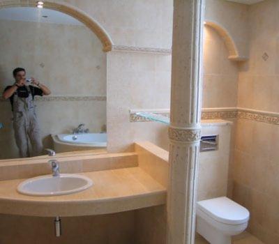 Wykończona łazienka w beżowych płytkach z dekoracyjnym lustrem łukowym, umywalką w blacie, toaletą i kolumną, odbicie fotografa w lustrze.