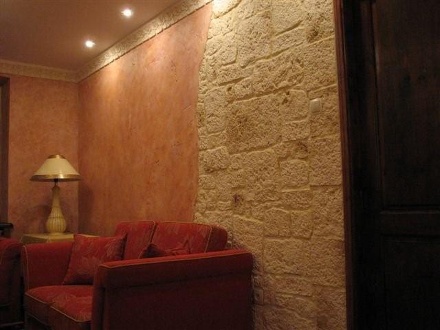 Fragment pokoju z dekoracyjną ścianą imitującą kamień, obok ściana w odcieniach pomarańczy z ozdobnym gzymsem, oraz czerwona sofa i lampka nocna.