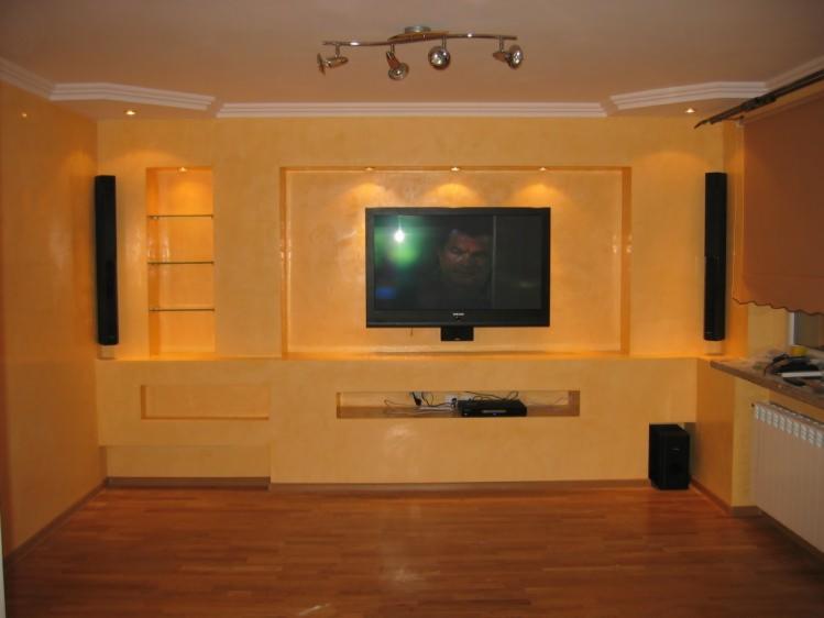 Żółta ściana z wnękami, podświetlane półki szklane, zamontowany telewizor, głośniki po bokach, parkiet na podłodze, oświetlenie sufitowe.