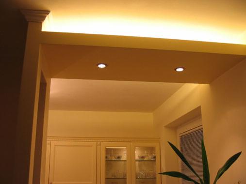 Oświetlenie sufitowe z dwoma punktowymi lampami w podwieszanym suficie, widok na przejście do innego pomieszczenia z zabudową meblową i rośliną doniczkową przy oknie.