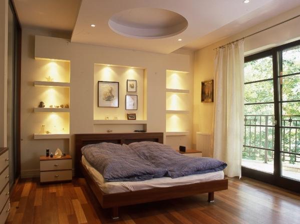 Elegancka sypialnia z drewnianą podłogą i łóżkiem, wnęki ścienne z oświetleniem punktowym i dekoracjami, okrągłe wgłębienie w suficie z oświetleniem i balkon z widokiem na zieleń.