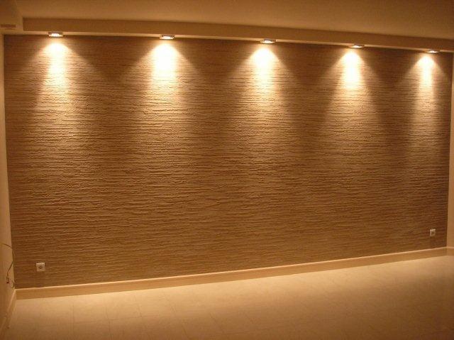 Teksturowana ściana w odcieniach beżu, oświetlona przez rząd punktowych lamp sufitowych, widoczne gniazdka elektryczne.