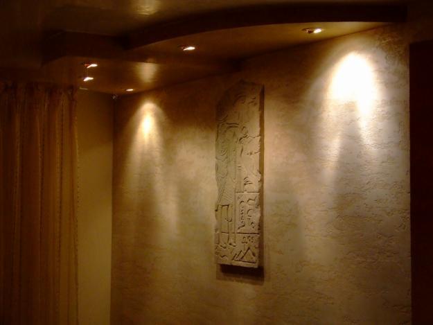 Pokój z dekoracyjną, teksturowaną ścianą w odcieniach beżu, podświetloną punktowymi lampami sufitowymi i zdobioną reliefem o motywach egipskich.