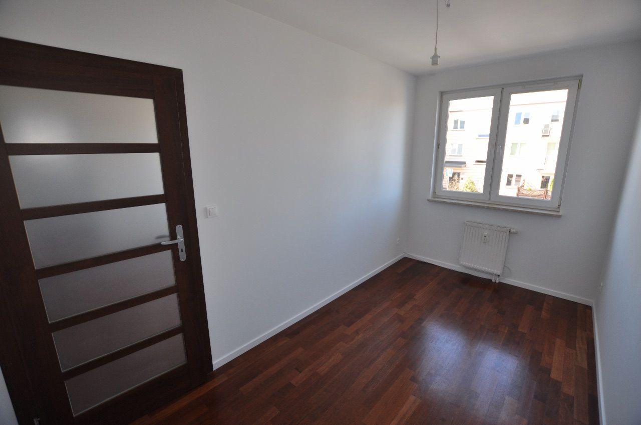 Pusty pokój po remoncie: ciemna drewniana podłoga, białe ściany, okno z widokiem na budynek, brązowe drzwi z matowym szkłem.