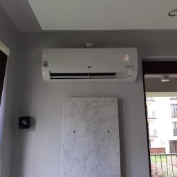 Nest Daniel Galas - Biały klimatyzator LG zamontowany na szarej ścianie nad dekoracyjną płytą z efektem marmuru w nowoczesnym wnętrzu z widokiem na balkon.