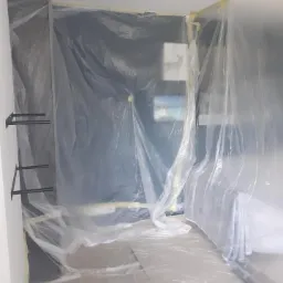 Przygotowanie pomieszczenia do prac remontowych: ściany i podłoga zabezpieczone folią malarską, widoczne uchwyty półek na ścianie i kabel odkurzacza na podłodze.