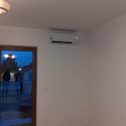 Nest Daniel Galas - Biała jednostka klimatyzacyjna zamontowana na ścianie w pokoju z widocznym przez okno balkonem i osobami na nim, podłoga zabezpieczona kartonem.