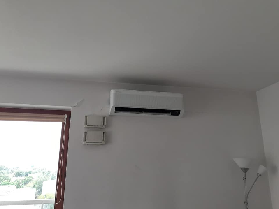 Biały klimatyzator ścienny zamontowany w pokoju z widocznymi śladami po poprzednim montażu, obok okna z roletą i lampą podłogową w tle.