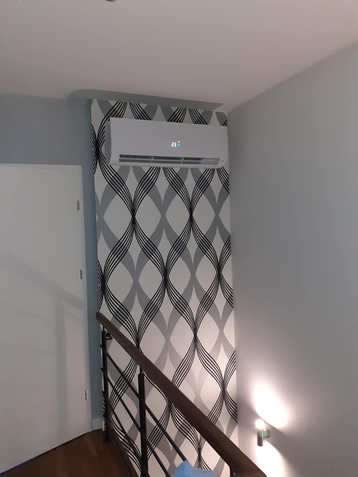 Biały klimatyzator ścienny zamontowany nad schodami z drewnianą balustradą i ozdobną tapetą w tle, wyświetlający temperaturę 23 stopnie.