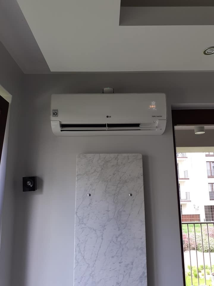 Biały klimatyzator LG zamontowany na szarej ścianie nad dekoracyjną płytą z efektem marmuru w nowoczesnym wnętrzu z widokiem na balkon.