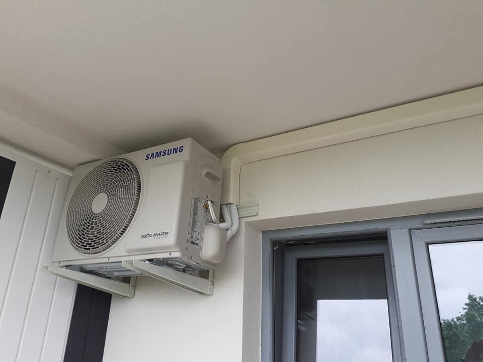 Biała jednostka zewnętrzna klimatyzatora Samsung Digital Inverter zamontowana na wspornikach na ścianie budynku, widoczne rury odprowadzające skropliny i przewody w białej listwie.