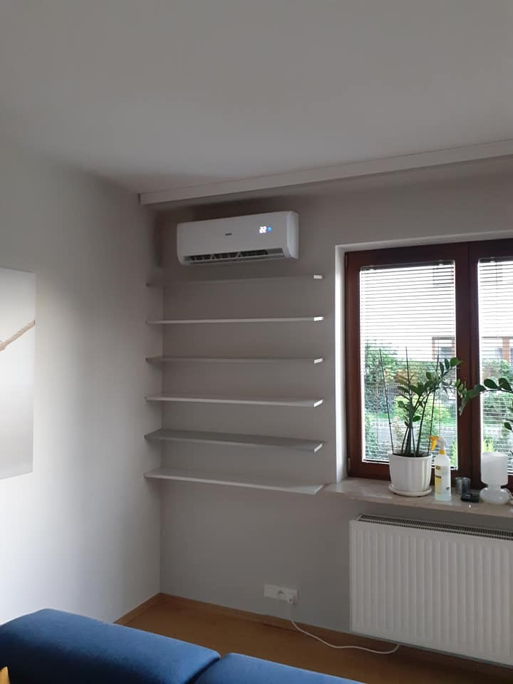 Biały klimatyzator ścienny zamontowany nad półkami w szarym pokoju, widoczne okno z roletami i grzejnik.