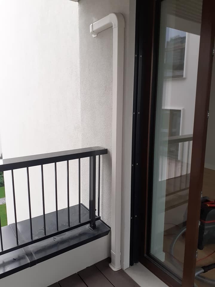 Biała rura odprowadzająca skropliny z klimatyzacji na balkonie nowoczesnego budynku, widoczna z zewnątrz, z czarną balustradą i przeszkleniem balkonu.