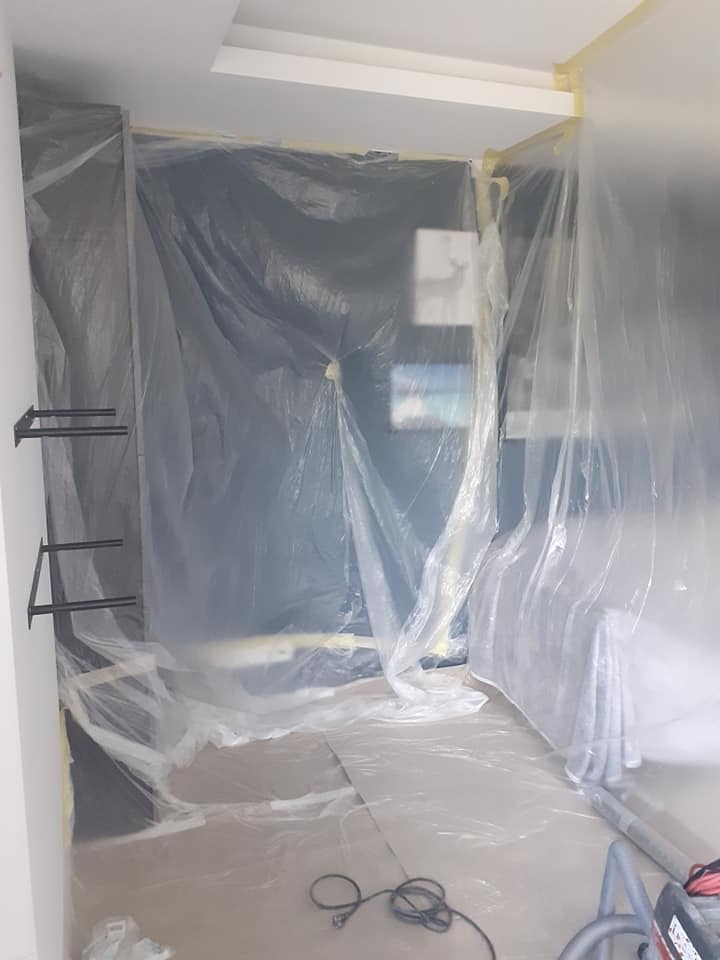 Przygotowanie pomieszczenia do prac remontowych: ściany i podłoga zabezpieczone folią malarską, widoczne uchwyty półek na ścianie i kabel odkurzacza na podłodze.