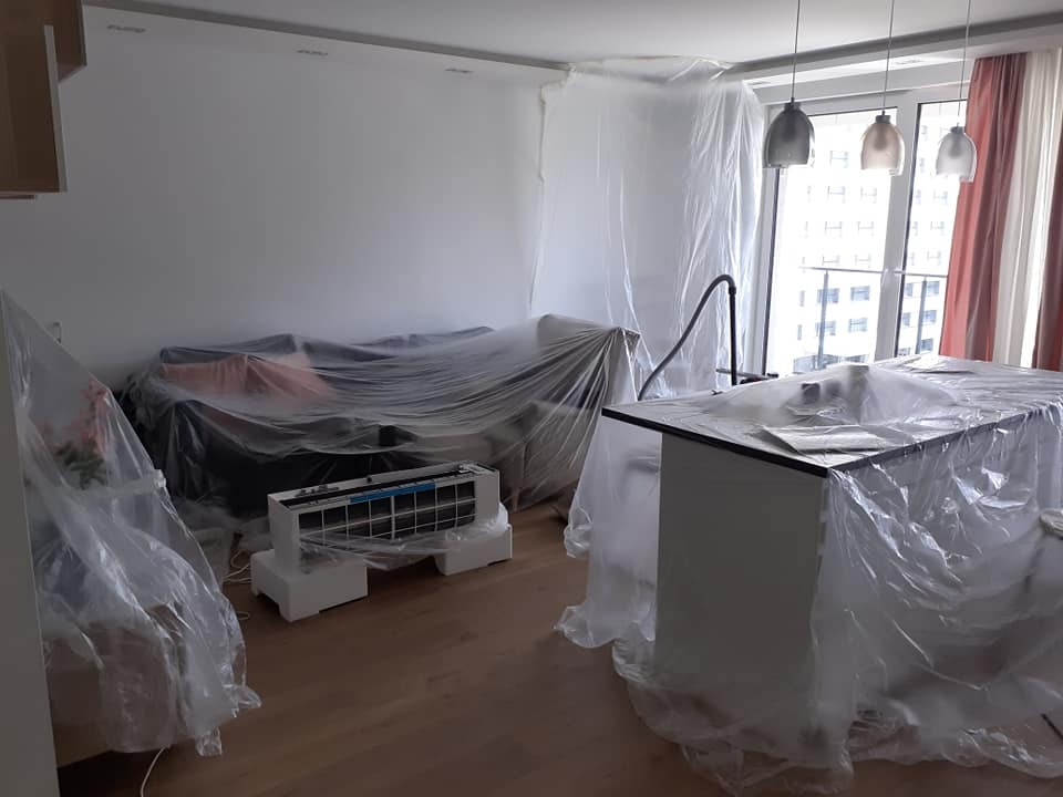 Salon w trakcie montażu klimatyzacji: meble zabezpieczone folią, jednostka wewnętrzna klimatyzatora na podłodze, widoczne okno z widokiem na miasto.