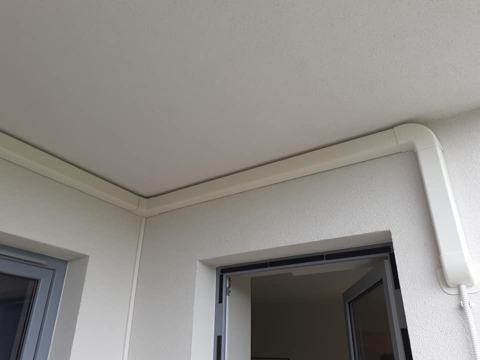 Biała listwa maskująca instalację biegnąca wzdłuż sufitu i ściany na balkonie, widoczny fragment okna i drzwi balkonowych.