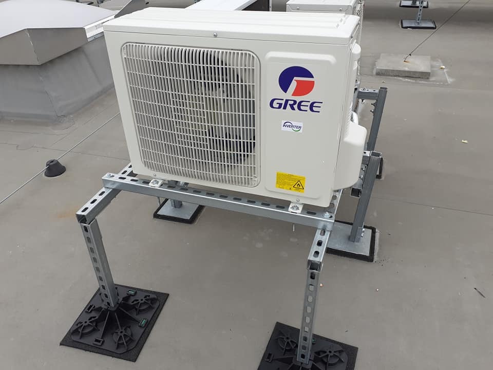 Biała jednostka zewnętrzna klimatyzacji marki Gree zamontowana na stalowej konstrukcji wsporczej na płaskim dachu.