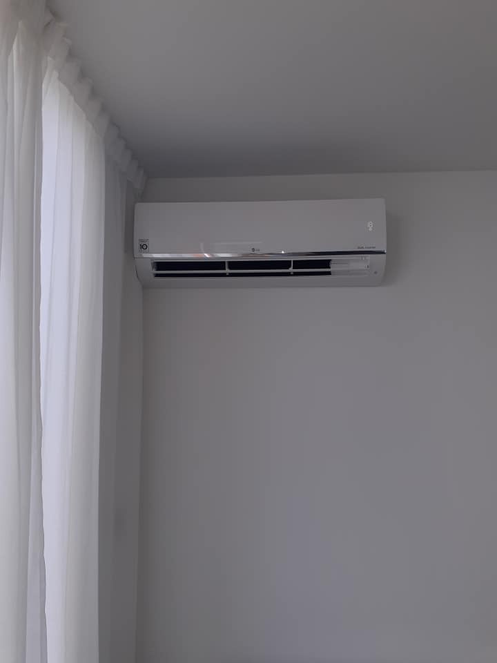 Biały klimatyzator ścienny marki LG zamontowany w pokoju z szarymi ścianami, obok zwiewnych białych zasłon