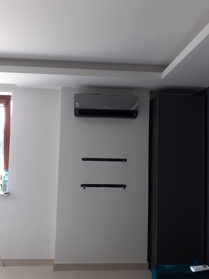 Szary klimatyzator LG zamontowany na białej ścianie w pokoju, poniżej widoczne uchwyty montażowe, obok ciemna szafa i okno z butelką wody.