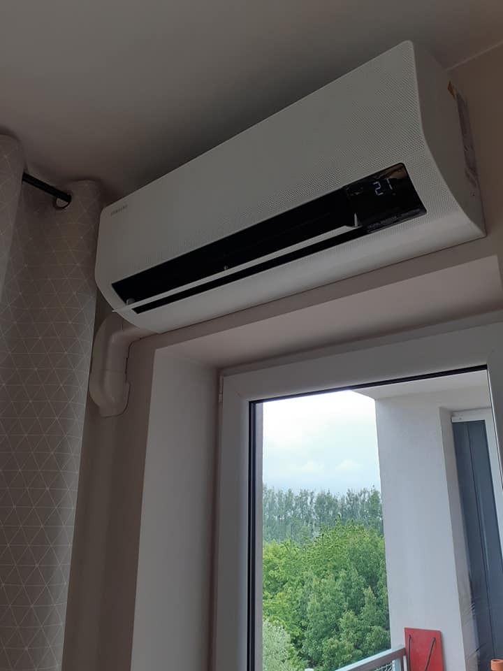 Biała klimatyzacja Samsung zamontowana nad oknem z widokiem na zieleń, widoczne rury instalacyjne i zasłona w geometryczne wzory.