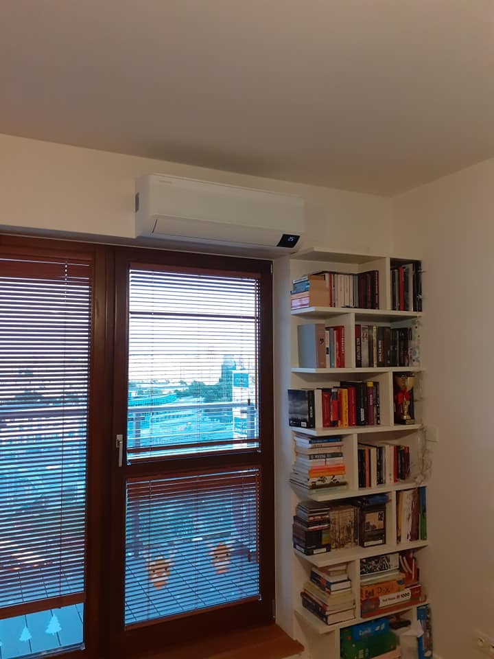 Biała jednostka klimatyzacyjna zamontowana nad oknem z żaluzjami w pokoju z półką na książki, widok na miasto w tle.