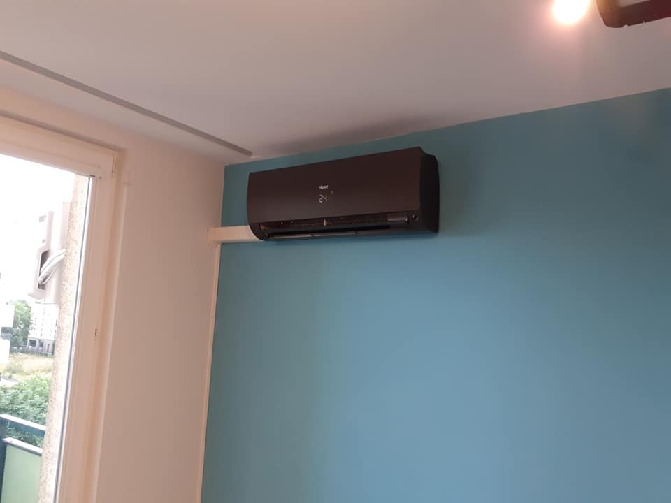 Czarny klimatyzator Haier zamontowany na błękitnej ścianie w pokoju, widok z boku, obok okno balkonowe.