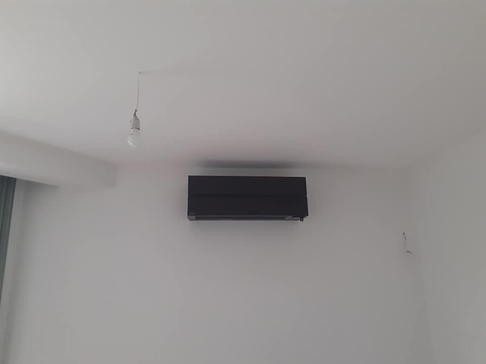 Czarny klimatyzator zamontowany na białej ścianie w pokoju, widoczna żarówka na kablu zwisająca z sufitu.