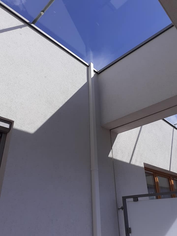 Szklany dach nad patio z widocznym niebem, białe ściany budynku z fakturą, cień padający na ścianę, widok z dołu.