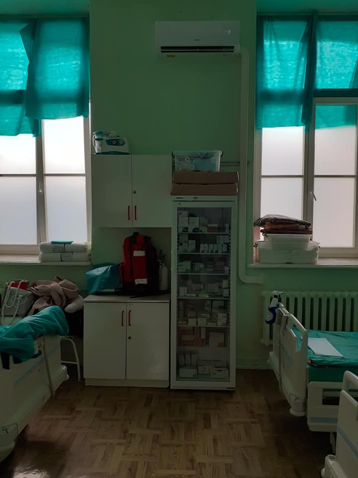 Wnętrze szpitalnej sali z dwoma łóżkami, szafkami i klimatyzatorem zamontowanym pod sufitem, widok na okna z turkusowymi zasłonami.