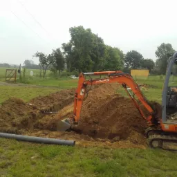 Pomarańczowa minikoparka Hitachi podczas wykonywania wykopu pod rurę kanalizacyjną na trawiastym terenie. Operator w kabinie, widoczne ułożone rury i świeżo wykopana ziemia.