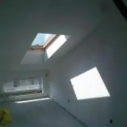 Białe wnętrze pokoju na poddaszu z dwoma oknami dachowymi, jedno z widoczną drewnianą ramą i wpadającym światłem, drugie zasłonięte folią malarską, ściany świeżo pomalowane na biało, widoczne ślady...