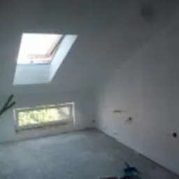 Poddasze po remoncie, widoczne okno dachowe i okno w ścianie szczytowej, świeżo pomalowane ściany na biało, wystające przewody elektryczne, surowa podłoga, brak mebli.