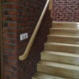 Drewniane schody z jasnymi stopniami i drewnianą balustradą, zlokalizowane w przestrzeni wykończonej cegłą w odcieniach czerwieni i brązu. Na ścianie widoczny włącznik światła.