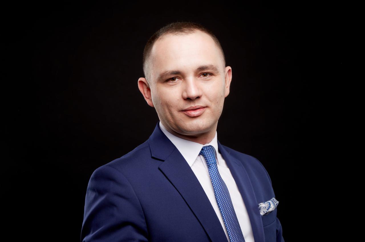 Portret mężczyzny w granatowym garniturze i błękitnym krawacie w białe kropki, z poszetką w tym samym odcieniu na czarnym tle.