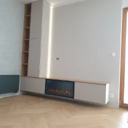 Nowoczesna szafa na wymiar z półkami i elektrycznym kominkiem w zabudowie, jasne drewno na podłodze ułożone w jodełkę, szara ściana z oświetleniem LED.