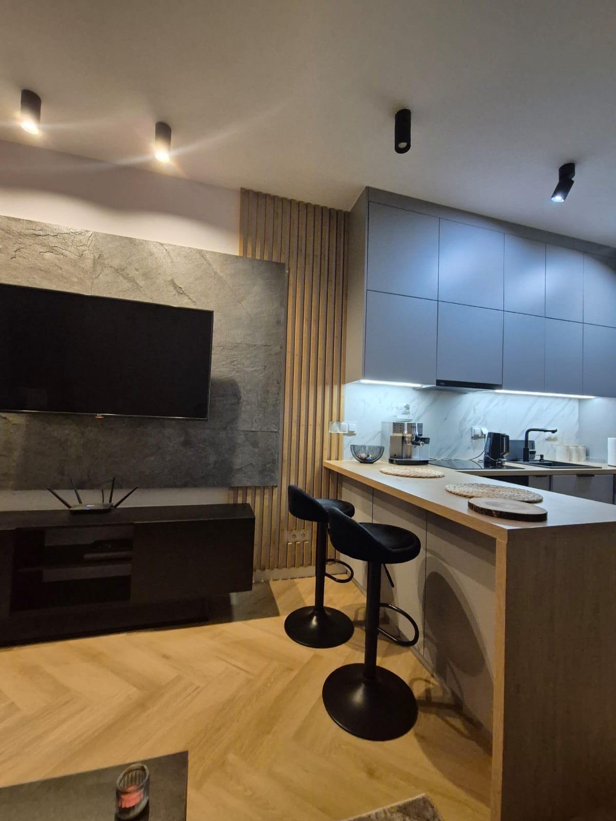 Nowoczesna kuchnia z szarymi szafkami, blatem barowym i dwoma czarnymi hokerami, oświetlona listwą LED pod szafkami wiszącymi, z widocznym ekspresem do kawy i czajnikiem, na tle ściany...