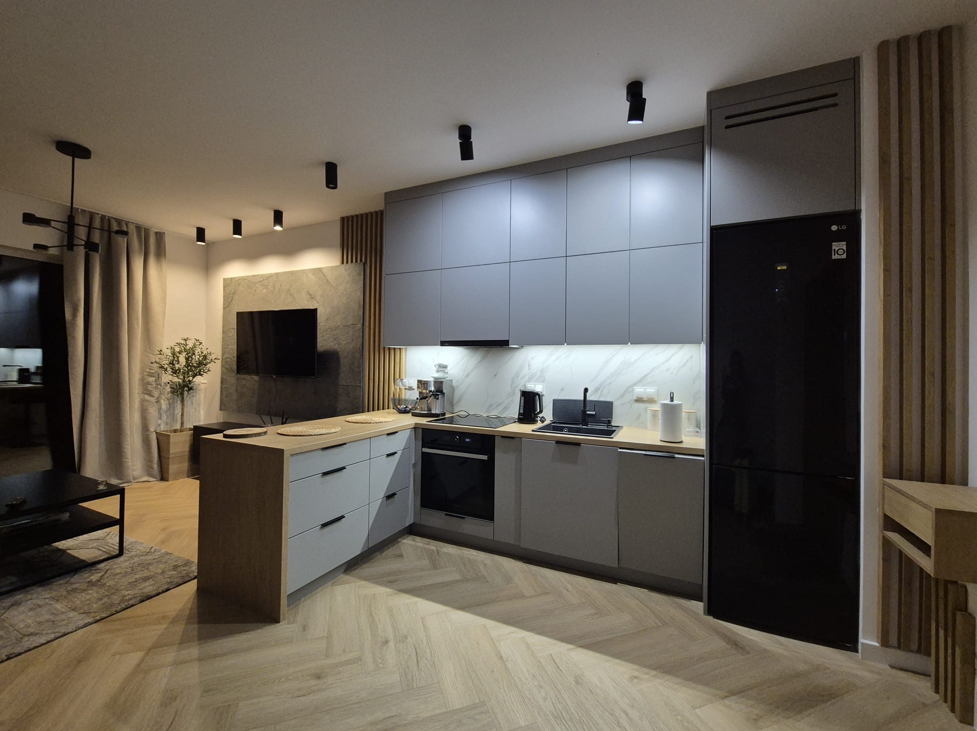 Nowoczesna kuchnia w odcieniach szarości z drewnianym akcentem, oświetleniem punktowym i czarną lodówką LG, widok na salon z telewizorem i roślinami.