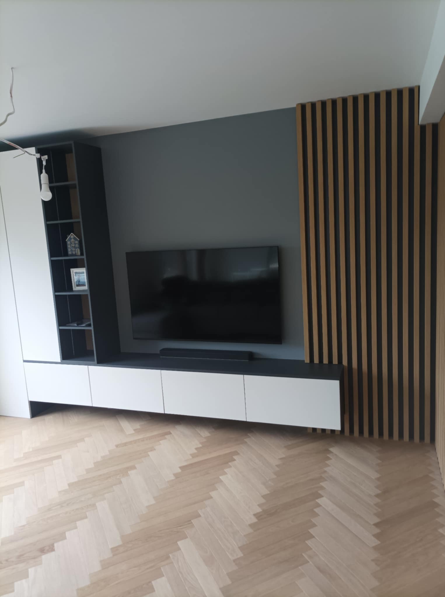 Nowoczesna szafka RTV na wymiar z białymi frontami i czarnymi półkami, uzupełniona o drewniane lamele na ścianie i parkiet w jodełkę. Minimalistyczny design.