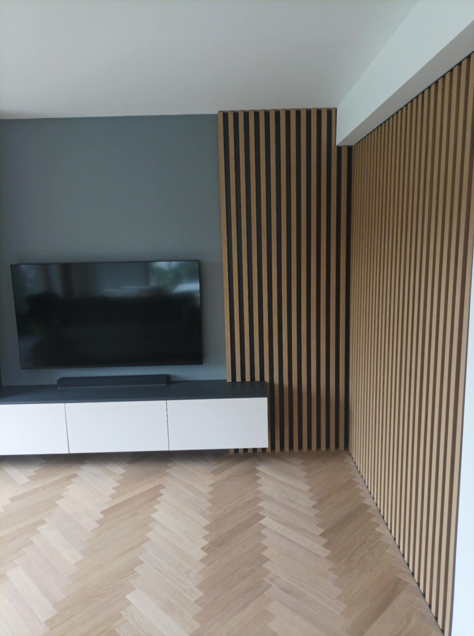 Nowoczesny salon z drewnianymi lamelami na ścianie, pod telewizorem biała szafka RTV, parkiet w jodełkę. Minimalistyczny design z akcentem naturalnego drewna.